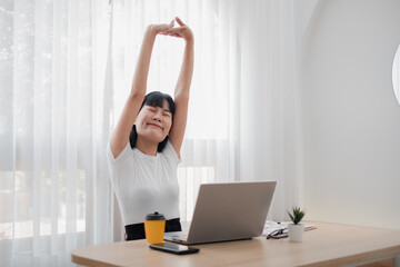 A young woman stretches at her desk with a laptop and coffee in a bright, modern home office.