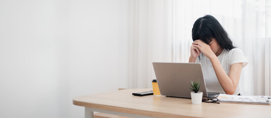 Woman feeling stressed while working from home, sitting at a wooden desk with a laptop and coffee...