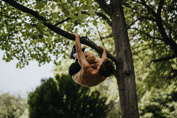 A shirtless athletic man demonstrates strength and agility by climbing a tree branch, surrounded by...