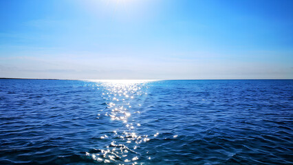 Obraz premium A single wave crests on a serene ocean, with a clear azure sky and sunlight casting glittering reflections on the water.