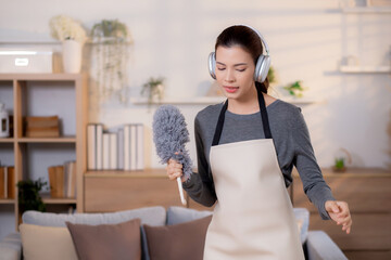 Young asian woman dusting while listening to music with headphones in a bright, organized living room, maid doing housework enjoying music and sing while dusting at home, lifestyle concept.