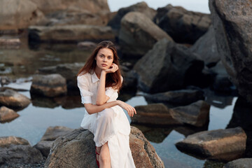 Serene woman in white dress contemplating by tranquil waters, hand on chin on rocky shore