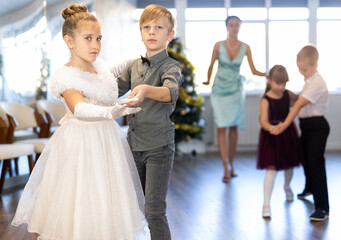 Obraz premium Positive elementary students in festive attire rehearsing ballroom dance number for upcoming Christmas party at school with interested female teacher in background..