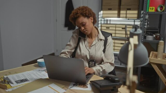 African american police woman on phone using laptop in an office at a detective's department.