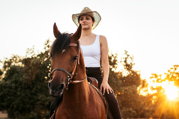 Young woman in hat riding a horse in the countryside at sunset
