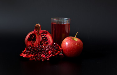 A glass of freshly squeezed fruit juice on a black background, next to pieces of a ripe red apple and a broken pomegranate fruit.