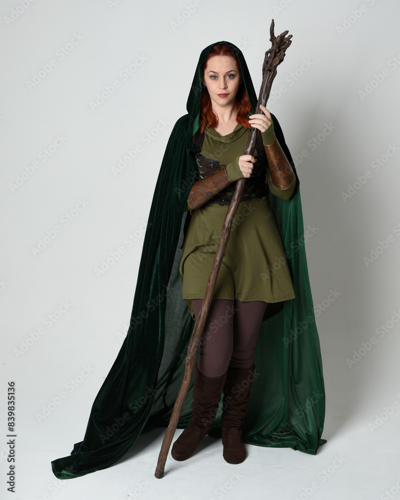 Sticker Full length portrait of red haired female model wearing green medieval fantasy costume leather armour, dramatic long flowing hooded velvet cape. Standing walking pose, isolated white studio background