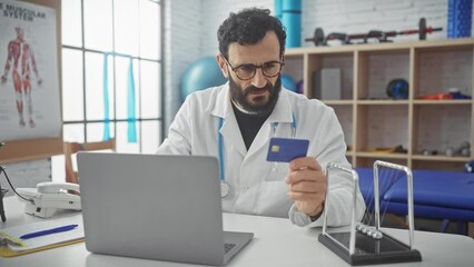 A bearded doctor in glasses examines a credit card at his laptop in a physiotherapy clinic office.