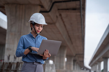 Asian female supervisor engineer working on her laptop under an overpass under construction,...