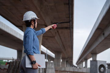 Fototapeta premium Asian female engineer supervisor under the overpass construction, using a laptop to analyze data and coordinate with her team, demonstrating leadership.