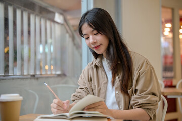 A young Asian female college student doing her homework at a cafe, writing something in a notebook.