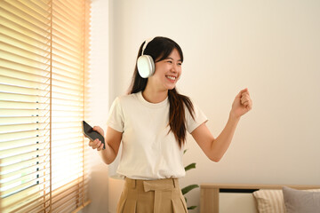 Cheerful young asian woman dancing while enjoying music through headphones in living room