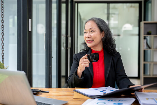 A mature Asian businesswoman is gazing at the view outside while sipping coffee at her desk. - Powered by Adobe