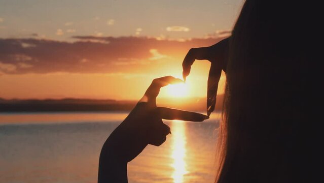 A girl on the shore of the lake creates a heart shape with her hands against the sunset background. A symbol of love and heart in the sun. The sun's rays.