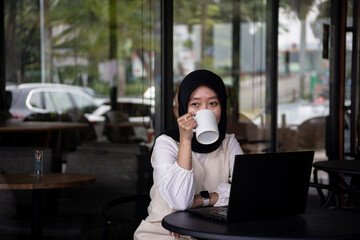 Young Indonesian woman wearing a black hijab sitting in a coffee shop. business woman working from coffee shop and drinking cup of coffee.