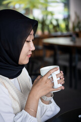 indonesian woman hijab sitting at coffee shop. business woman work from coffee shop and holding cup...