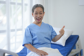 Happy elderly patient giving thumbs up while sitting on hospital bed, showing positive healthcare...