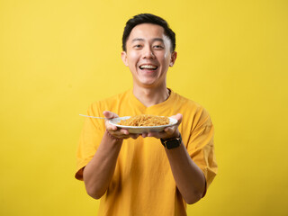 A young man in a yellow shirt is enthusiastically eating a plate of noodles with a fork against a...