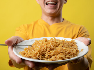 A young man in a yellow shirt is enthusiastically eating a plate of noodles with a fork against a...