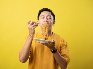 A young man in a yellow shirt is enthusiastically eating a plate of noodles with a fork against a...