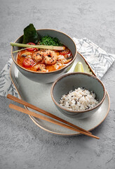A popular dish of Asian cuisine is Tom Yam soup, cooked with shrimp, lemongrass, galangal and lime in a bowl on a gray background. Spicy an hot Thai soup is served with boiled white rice.