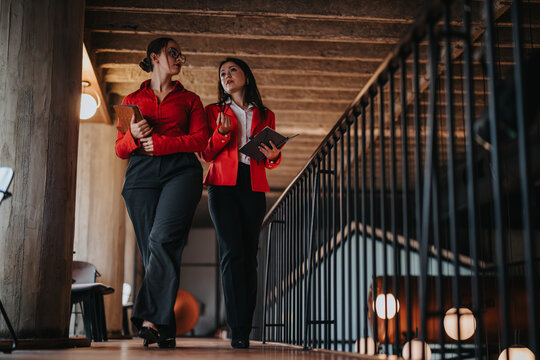 Two young business entrepreneurs walking and discussing ideas in a modern office environment. Both are wearing professional attire and holding notebooks.