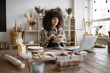 Female pottery artist shaping clay on wheel while referencing laptop in cozy home studio. Creative,...