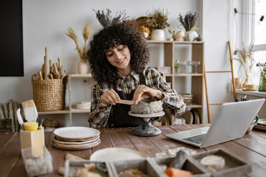 Young woman sculpting clay in cozy home studio with laptop on table. Artistry, concentration, and creativity in a modern crafts workspace.