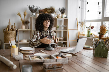Young woman working on pottery while using laptop in creative studio. Artist shaping clay and referencing design on computer.