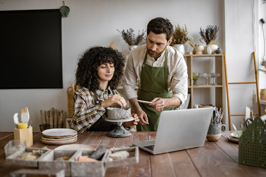 Man instructing woman on pottery techniques in studio. Collaborative learning environment with laptop and art supplies.