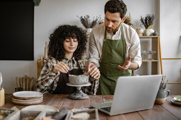 Male pottery instructor teaching female student in studio using laptop. Woman shaping clay with...