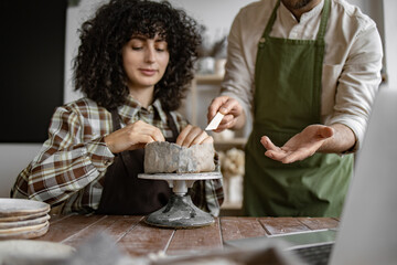 Female pottery student learning ceramic techniques from male instructor in craft studio. Hands-on pottery lesson and creative workshop.