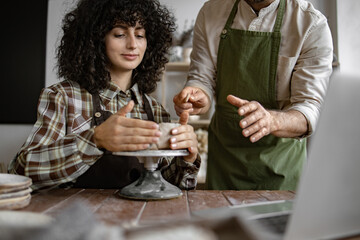 Man instructing woman in shaping clay on potter's wheel in cozy studio. Creative activity and crafting.