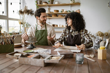 Bearded mature man in apron sculpting clay vase while woman uses rolling pin in pottery studio. Creative expression and teamwork.