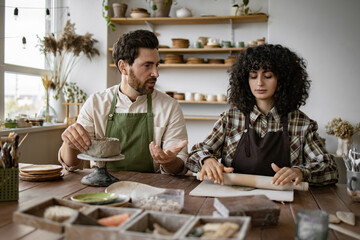 Bearded mature man and woman making pottery together in cozy workshop. Man sculpts vase, woman uses rolling pin for clay.