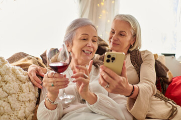 Two middle-aged women relax in a camping van, enjoying a glass of wine and checking smartphone