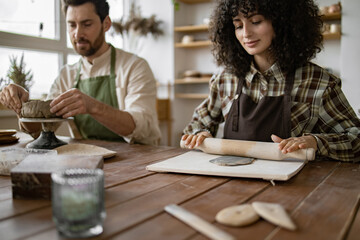 Bearded mature man and woman creating clay art at pottery studio. Man sculpts a vase, woman rolls clay with a rolling pin.
