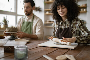 Bearded mature man and woman sculpting in a pottery class. He creates a vase, while she rolls clay with a rolling pin.