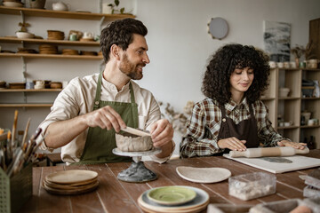 Bearded mature man in apron sculpts vase while woman rolls clay with rolling pin during pottery...