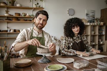 Bearded mature man in apron sculpting vase and woman rolling clay with rolling pin in pottery studio. Creative artistic process.