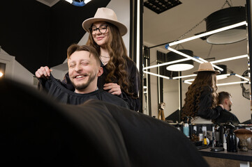 Obraz premium A young man with a beard in a black cape sits on a chair in a barbershop. The client is waiting for a haircut of the beard.