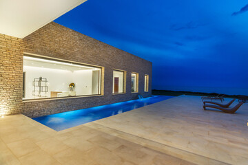 Modern House With Swimming Pool at Night.
A modern house with glass walls and a flat roof is illuminated from within, casting a warm glow across an adjacent swimming pool. The pool reflects the night 