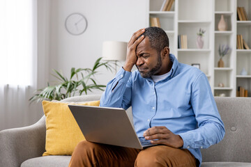 Frustrated man sitting on the couch with a laptop, holding his head. He appears to be dealing with...