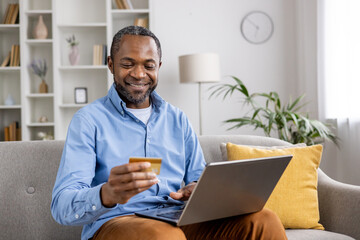 Smiling man holding a credit card and using a laptop while shopping online at home. Comfortable and modern living environment