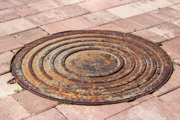 A rusty manhole cover is placed on a brick sidewalk in an urban setting