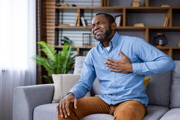 Man holding chest, experiencing pain while sitting on couch in comfortable living room setting, depicting heart attack or chest pain symptoms