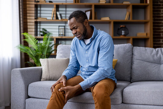 Man holding his knee in pain while sitting on a couch. The background shows a stylish living room with modern decor.