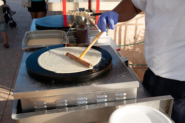 A man is making a dessert on a pan. The pan is on a table and there are other pans and bowls on the table. The man is wearing a blue shirt and a white apron