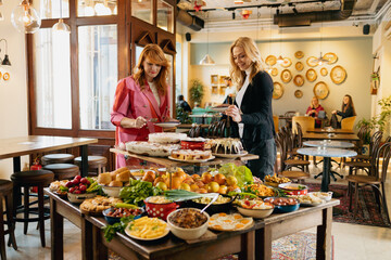 Two woman standing in front of delicious food buffet assorted on wooden table in restaurant.