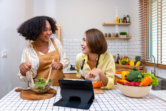 African American woman and her asian friend is helping each other preparing healthy food with variety of vegetables while looking at the cooking instruction from internet by using digital tablet